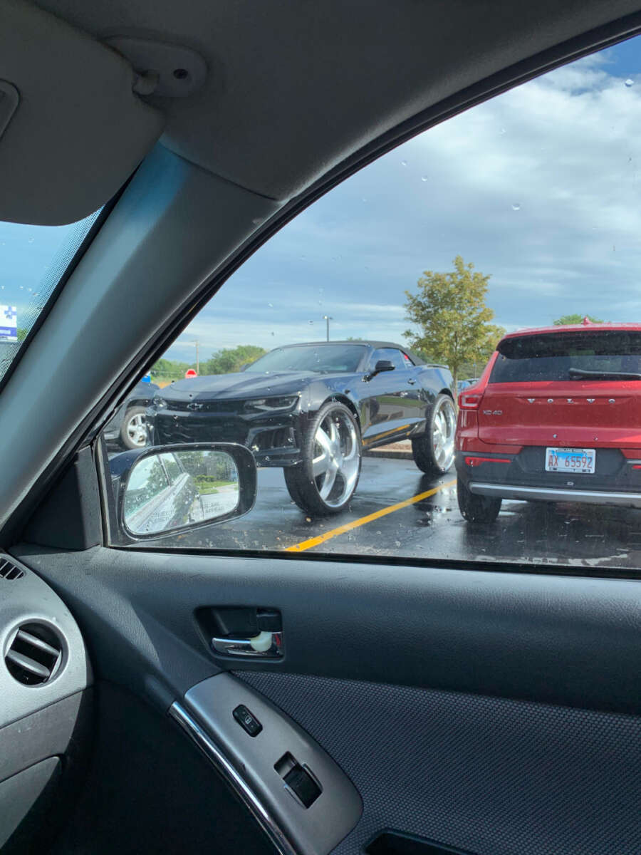 A shiny black sports car with oversized chrome wheels is parked next to a red SUV in a wet parking lot, viewed from inside another vehicle through a rainy window.