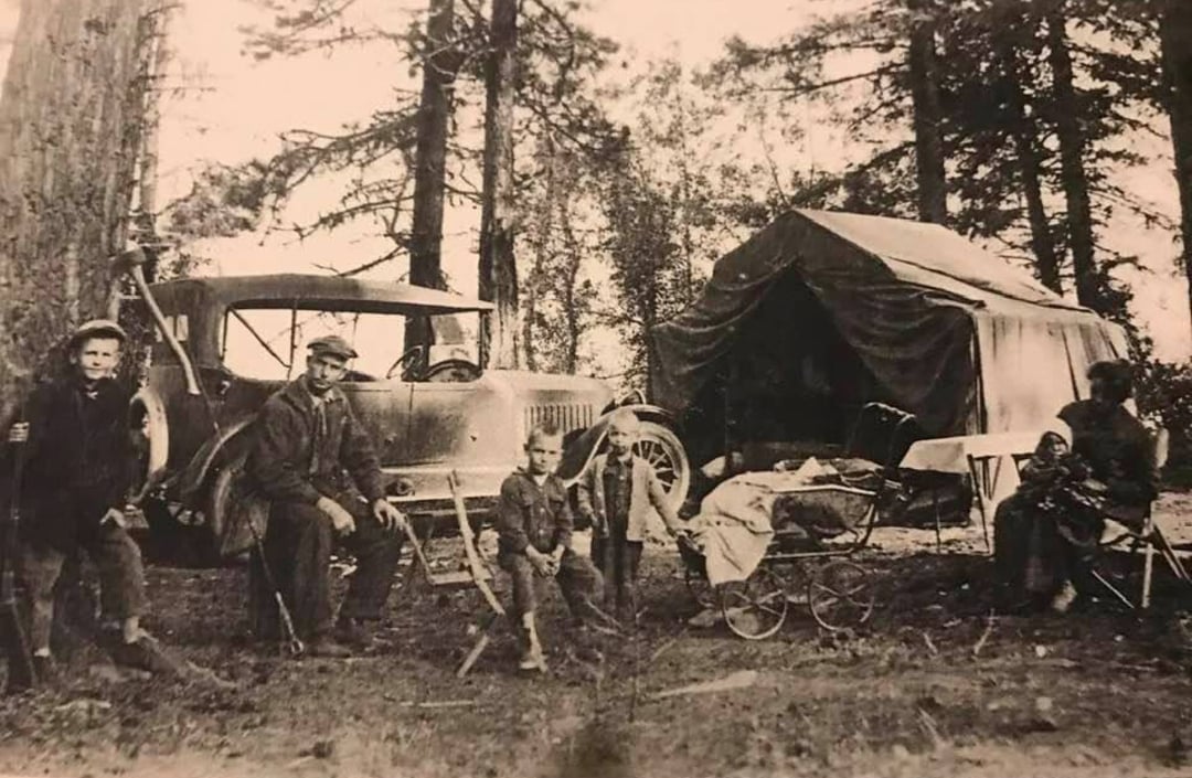 A black-and-white photo of a family camping in the woods with a vintage car, a canvas tent, and camping furniture. Several adults and children pose among tall trees, with camping gear and a stroller visible.