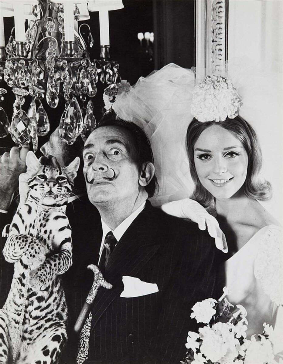 A man in a suit with a mustache and cane poses with a woman in a wedding dress and gloves, both smiling beside a chandelier. The man holds an ocelot, which looks directly at the camera.