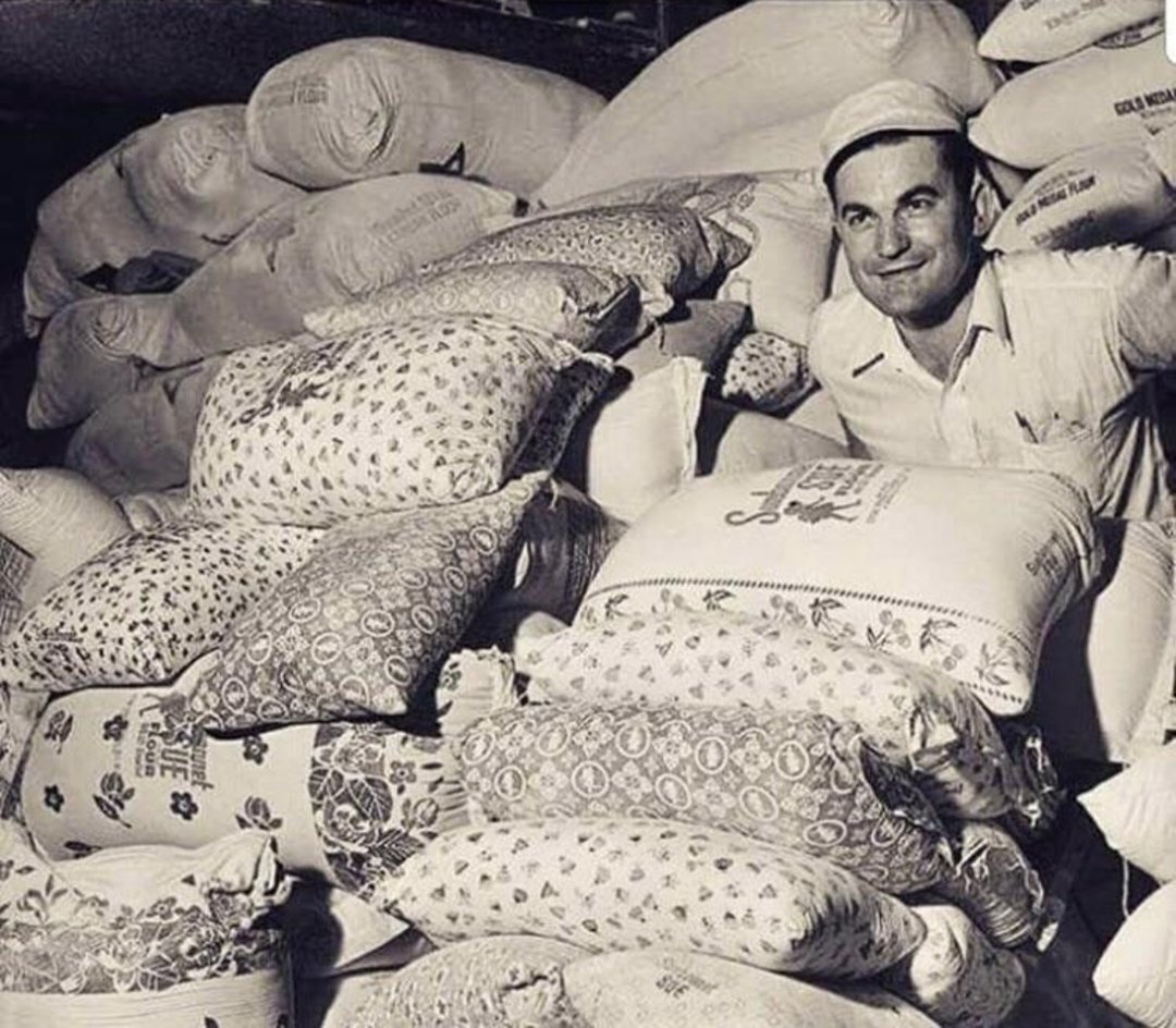 A man in a white shirt and cap stands among a large pile of patterned flour sacks, smiling. The scene appears to be in a storage or warehouse area, with many sacks stacked around him.