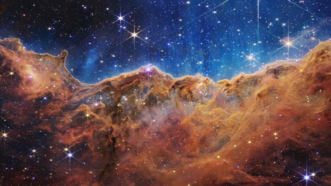 A vibrant image of a cosmic landscape shows bright stars scattered across a blue and black space background, with a jagged, orange-brown nebula cloud stretching horizontally in the foreground.