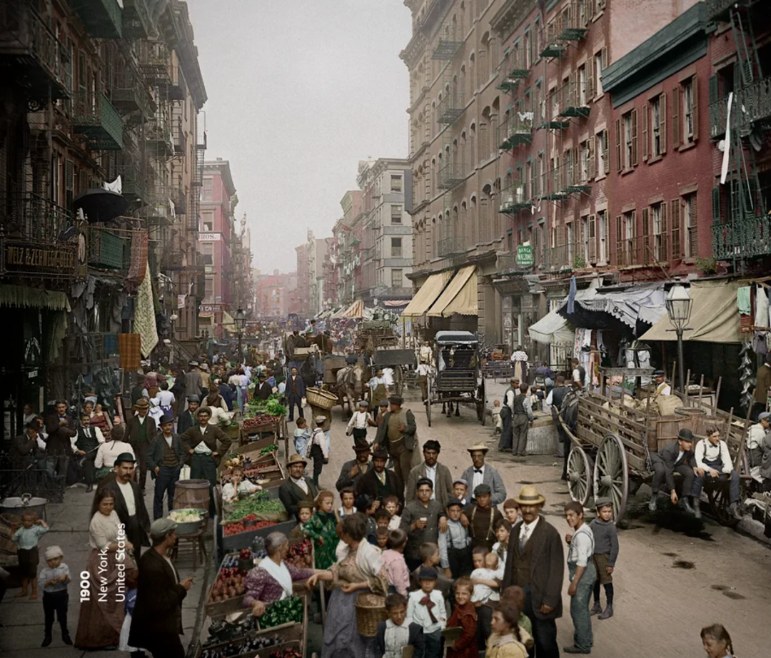 A bustling New York City street market in 1900, crowded with people, horse-drawn carts, vendors selling produce, and tall brick buildings lining both sides of the busy street.