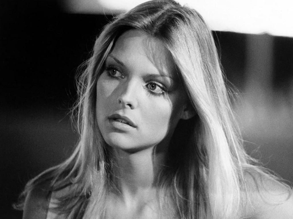 A black-and-white photo of a young woman with long, straight blonde hair, looking slightly to the side with a thoughtful expression. She is lit by soft light, and the background is blurred.