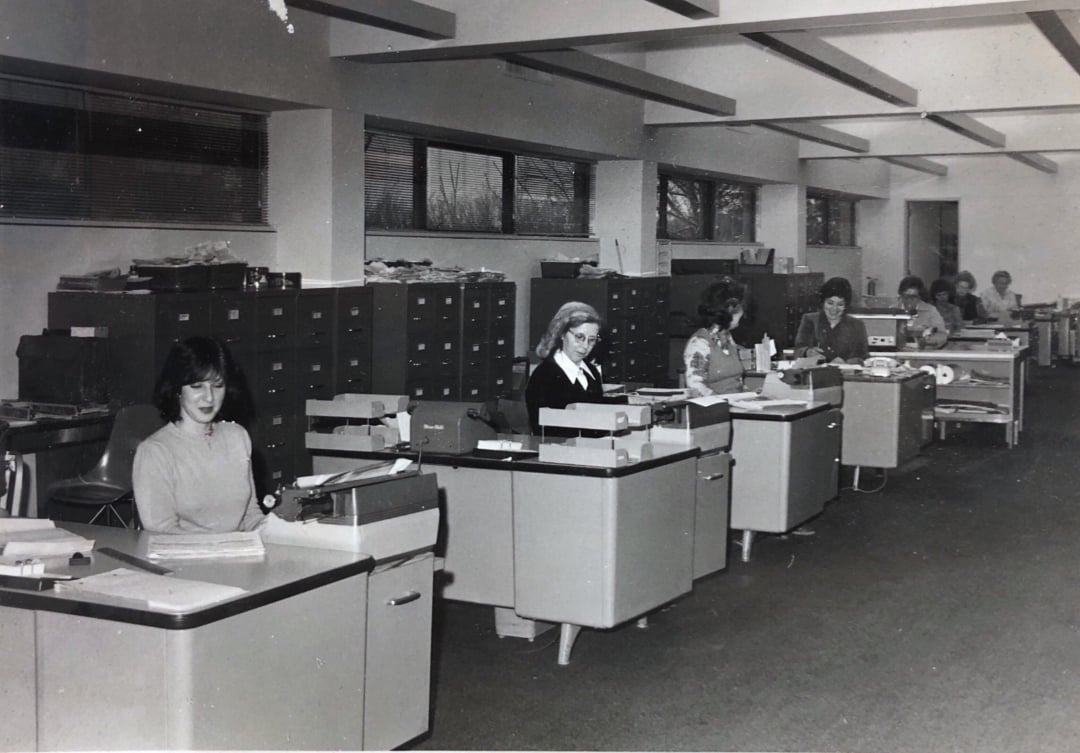 Black and white photo of women working at typewriters in a row of desks in an office; filing cabinets line the wall, and large windows let in natural light. The setting appears to be from the mid-20th century.