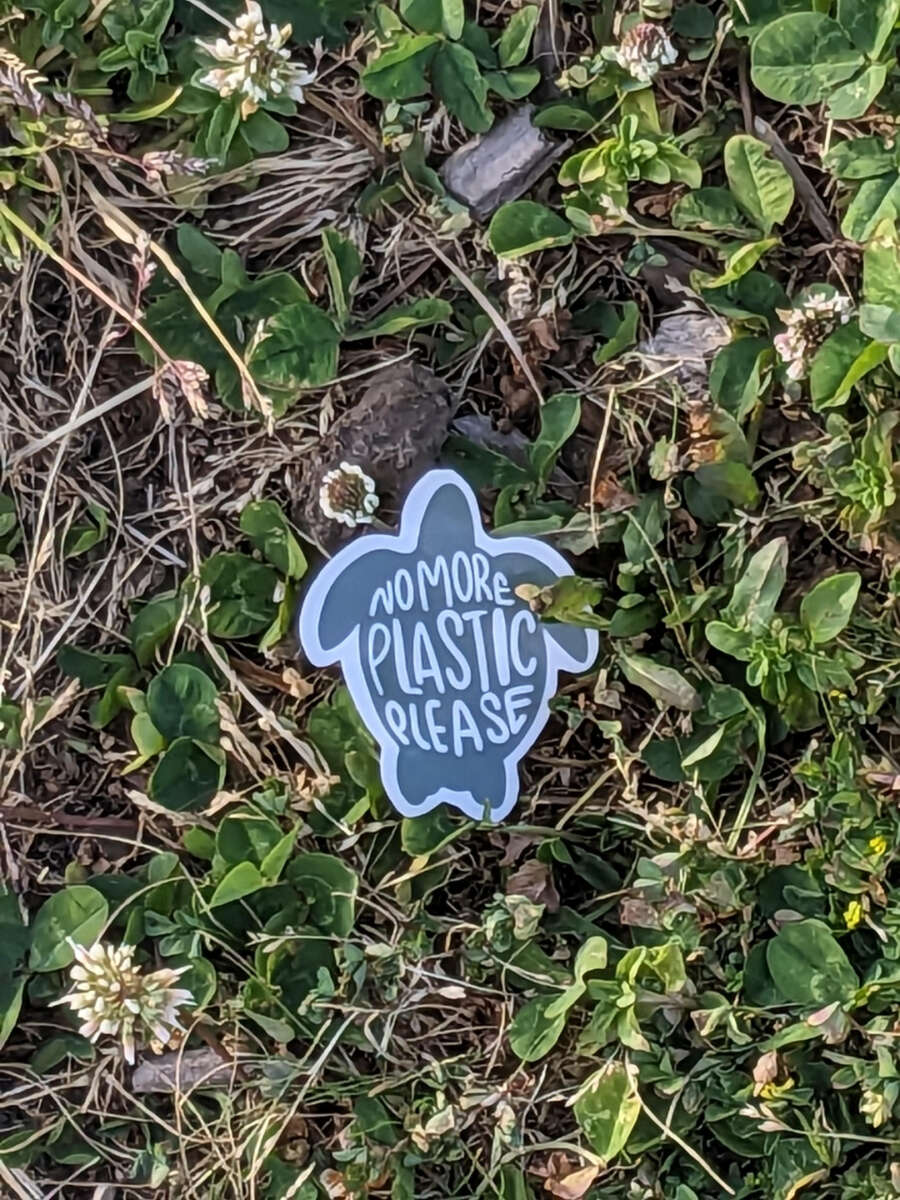 A sticker shaped like a turtle on grass and clover reads "No More Plastic Please" in handwritten letters.