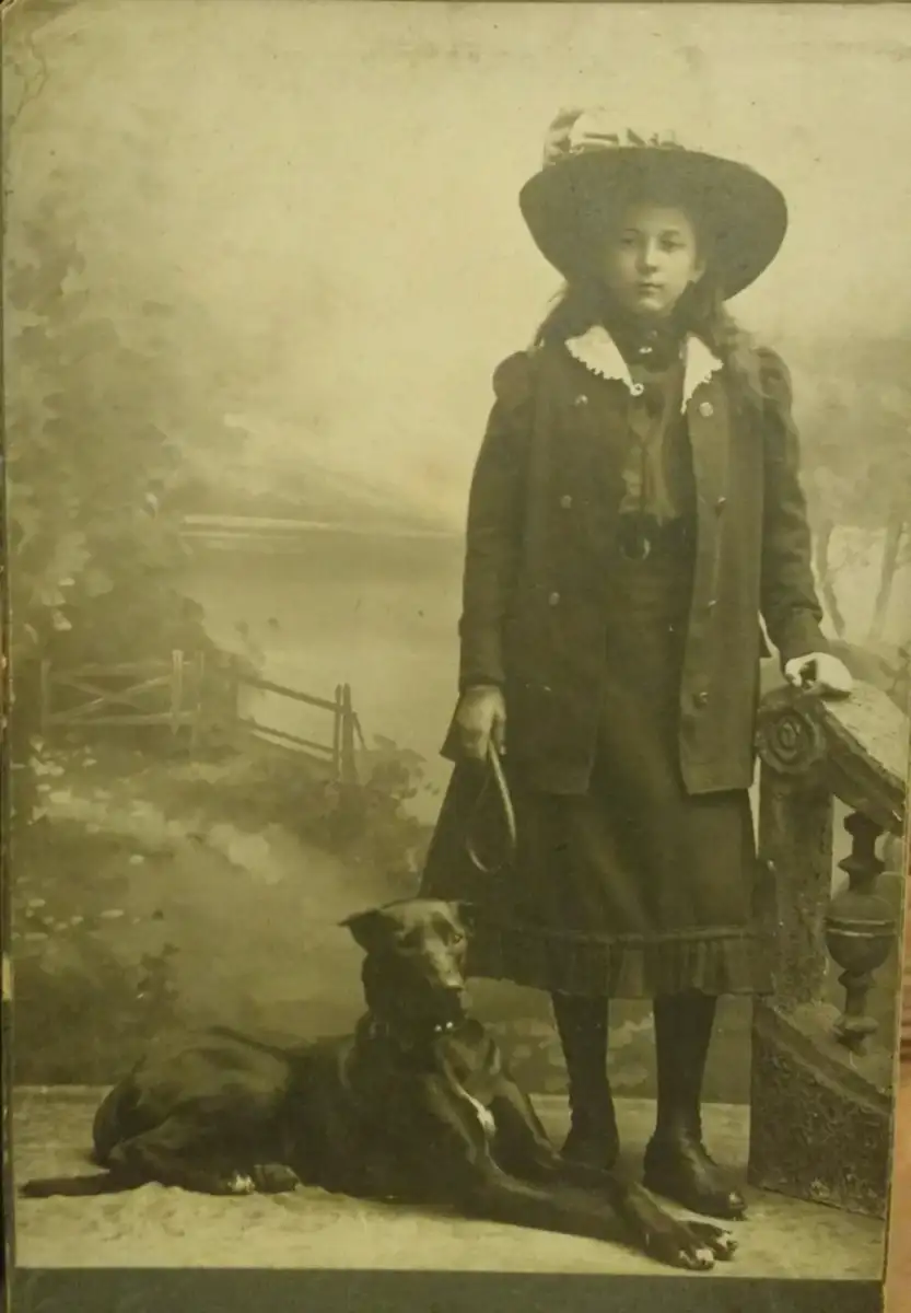 A young girl in old-fashioned clothing and a large hat stands on a stone step holding a leash, with a large dog lying at her feet; a painted countryside backdrop is behind them.