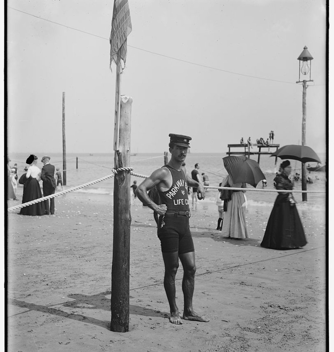 A male lifeguard in a uniform and cap stands on a beach near a rope barrier, with people in old-fashioned clothing and umbrellas behind him, and a pier and ocean in the background.