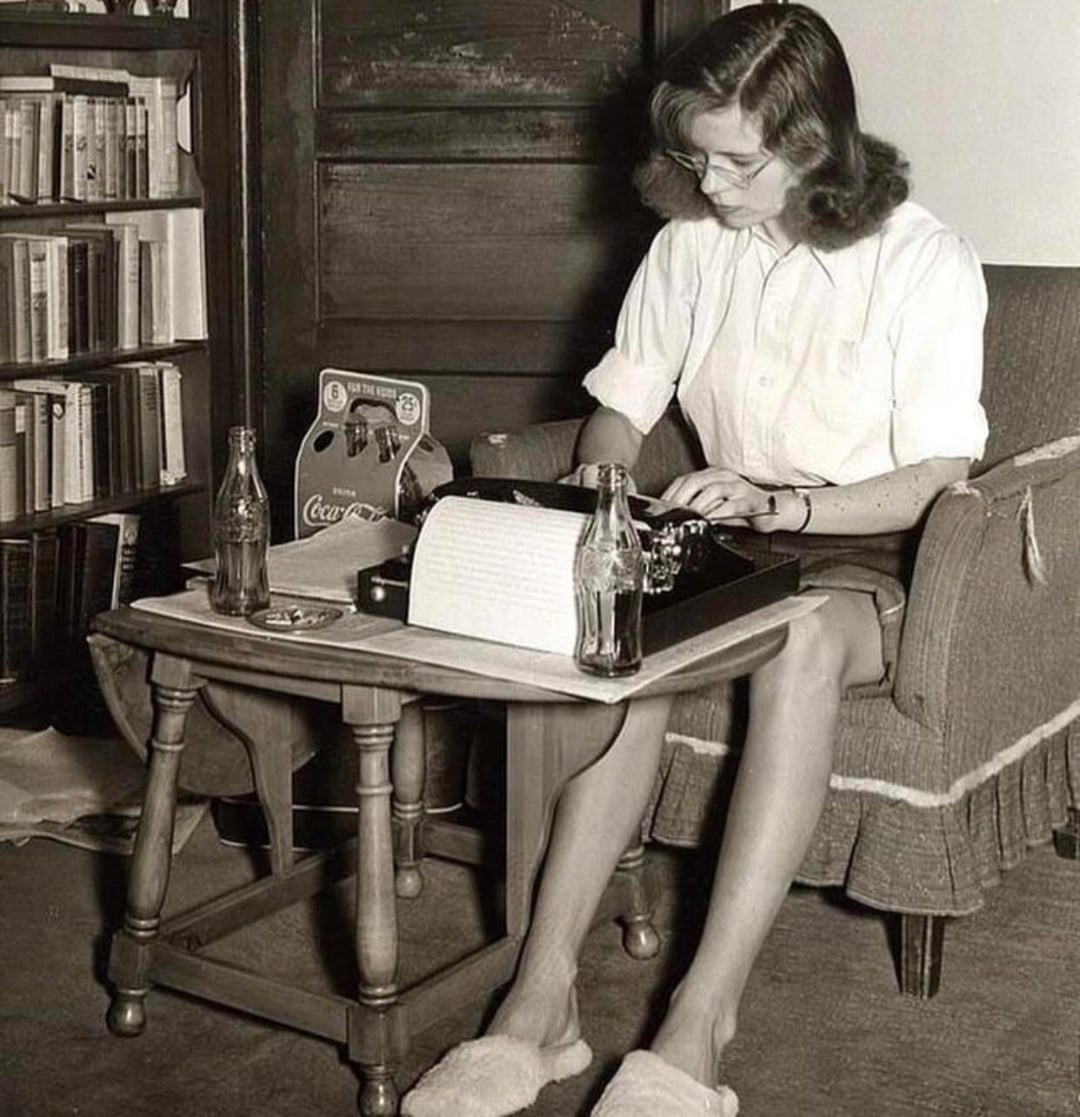 A woman wearing glasses and slippers sits in an armchair typing on a typewriter. A table in front of her holds a bottle of Coca-Cola, papers, and a Coca-Cola carton. Bookshelves and a closed door are in the background.