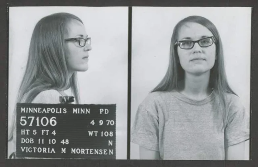 A black-and-white mugshot shows a young woman with long hair and glasses, photographed from the side and front. The placard reads: “Minneapolis Minn PD 57106, 4 9 70, HT 5 FT 4, WT 108, DOB 11 10 48, Victoria M Mortensen.”