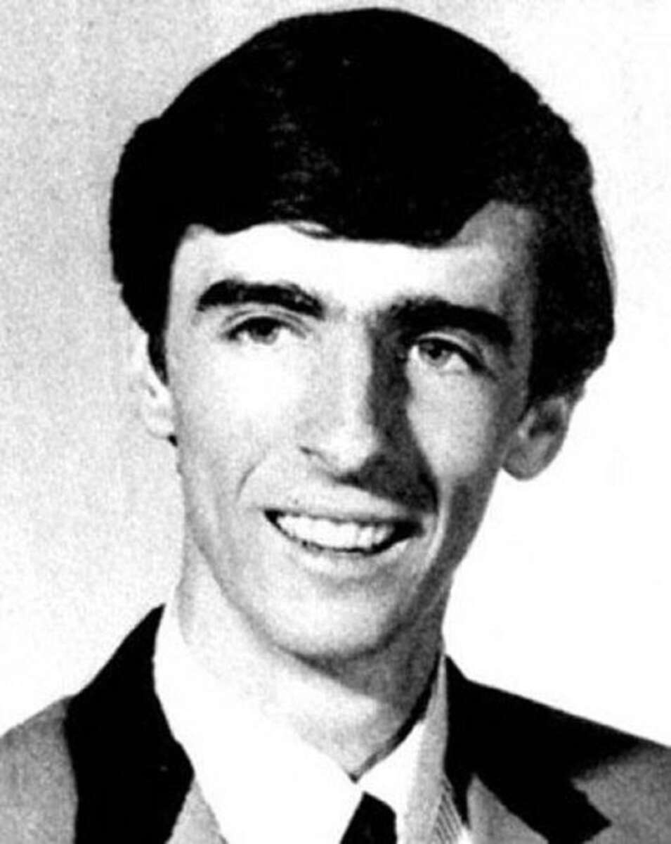 A black and white portrait of a young man with dark hair, wearing a suit jacket, dress shirt, and tie, smiling at the camera.