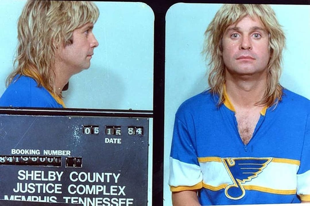 A man with shaggy blonde hair wearing a blue and yellow hockey jersey is photographed for a mugshot. The sign reads "Shelby County Justice Complex, Memphis Tennessee" with the date 05-15-84.