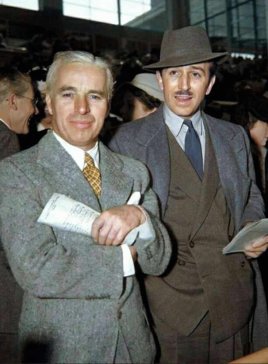 Two men in vintage suits and ties stand indoors, one holding papers and smiling, the other wearing a hat and mustache. Both appear at a formal or public event with other people visible in the background.