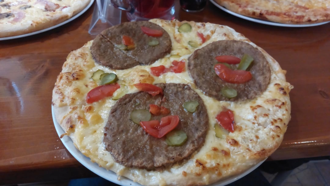 A pizza topped with three large hamburger patties, sliced pickles, and red bell pepper pieces, on a cheesy crust, served on a white plate on a wooden table. Another pizza is partially visible in the background.