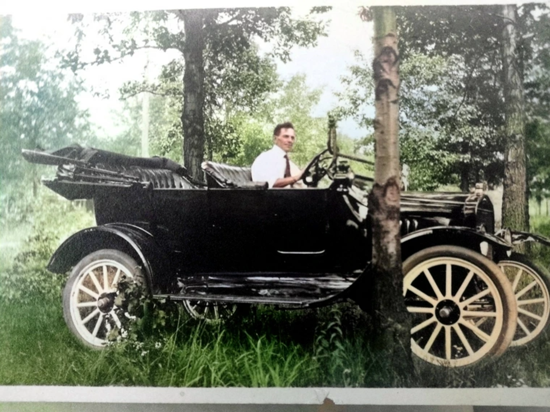 A man in a white shirt and tie sits in a vintage black car with white wheels, parked on grass among trees in a lush, green outdoor setting.