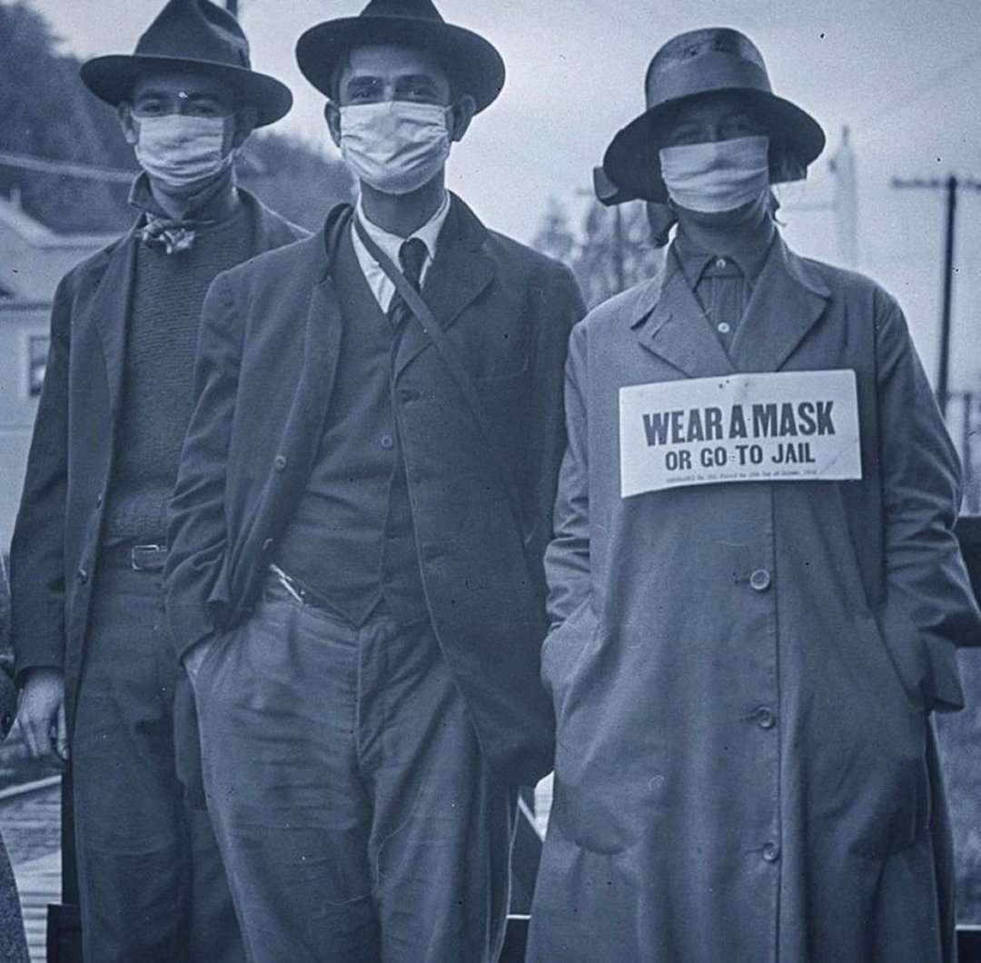 Three people wearing face masks and hats stand outdoors. One person in front wears a coat with a sign that reads, "WEAR A MASK OR GO TO JAIL." The image is in black and white, suggesting a historical context.