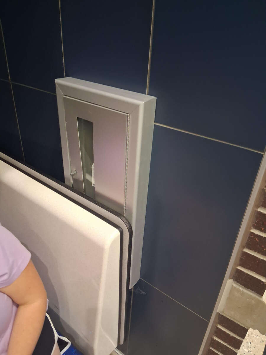 A metallic baby changing table mounted on a dark tiled wall in a restroom, shown in the closed upright position. Part of a person’s arm and pastel shirt are visible on the left side of the image.