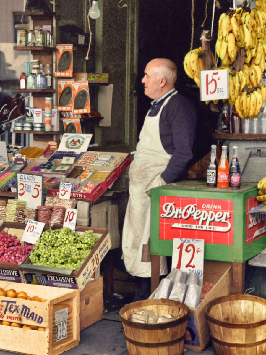 A man in a white apron stands in a colorful market stall with bananas, grapes, candy, soda bottles, and produce, surrounded by price signs and shelves of goods.