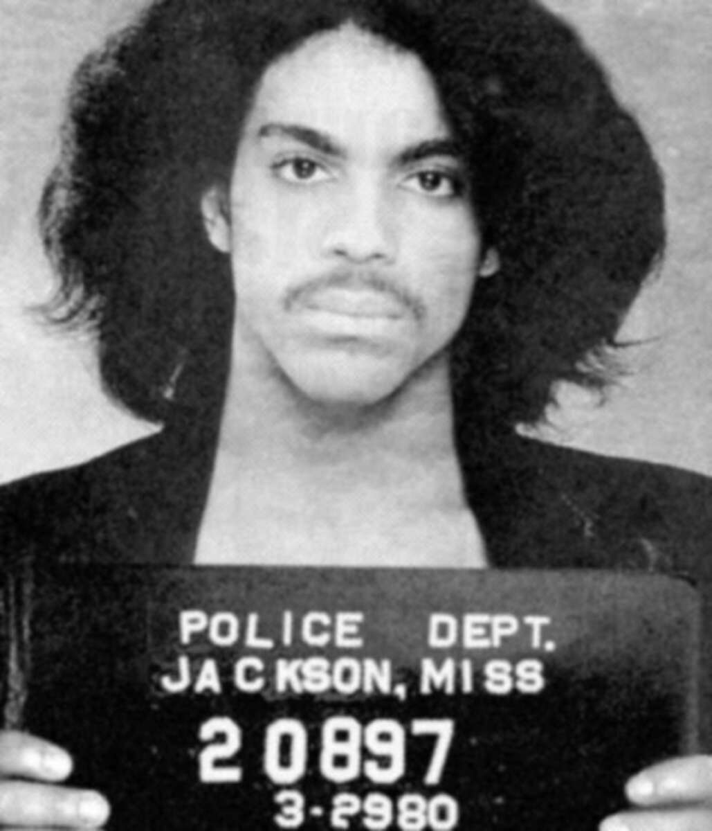 A person with medium-length, wavy hair holds a police department sign in front of their chest for a mugshot, with "Jackson, Miss" and identification numbers visible on the sign.