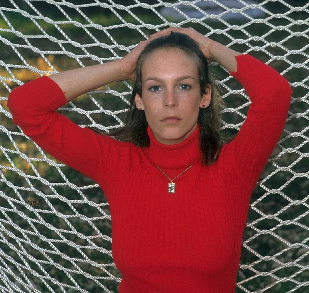 A woman in a red turtleneck sweater stands with her hands on her head, posing in front of a netted hammock outdoors with blurred greenery in the background.