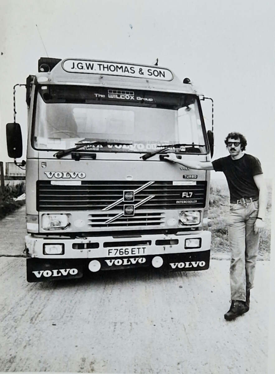 A man wearing sunglasses and a dark shirt leans against a large Volvo truck labeled "J.G.W. Thomas & Son" on a rural road, smiling at the camera. The photo is in black and white.