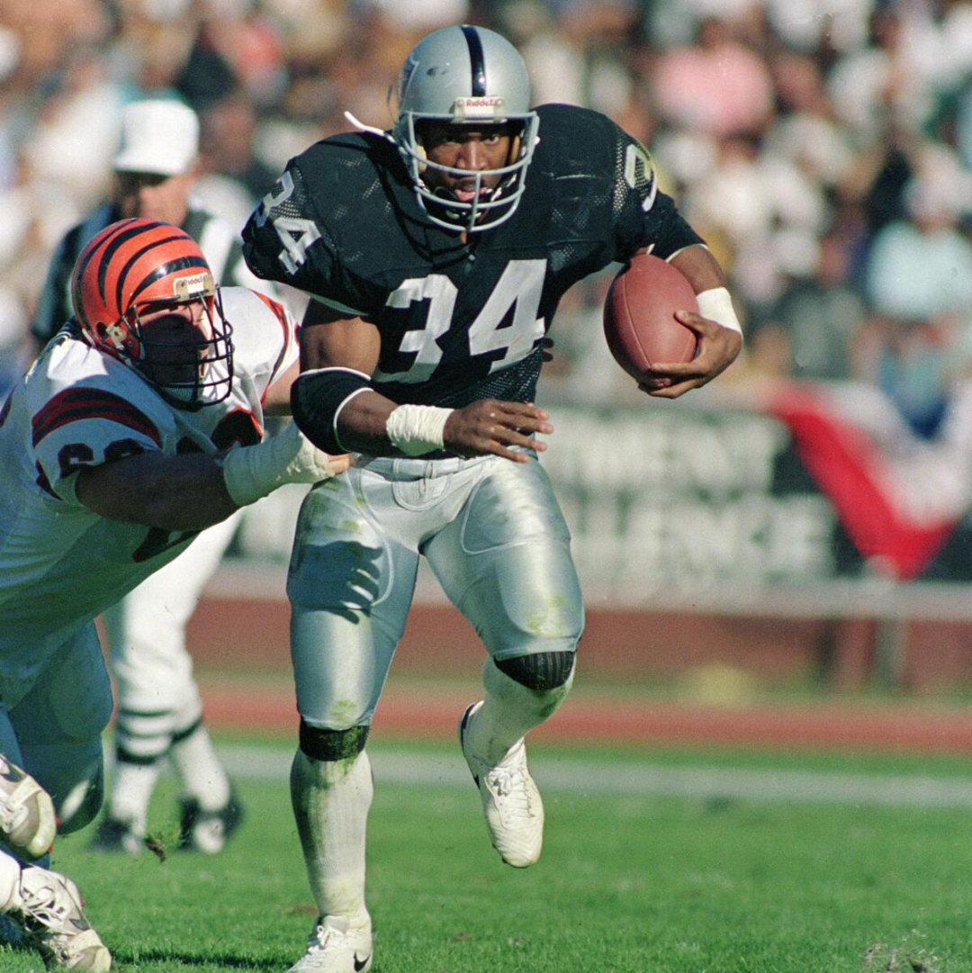 A football player in a black jersey with the number 34 runs with the ball as a defender in a white and orange uniform attempts to tackle him during a game on a sunny day.