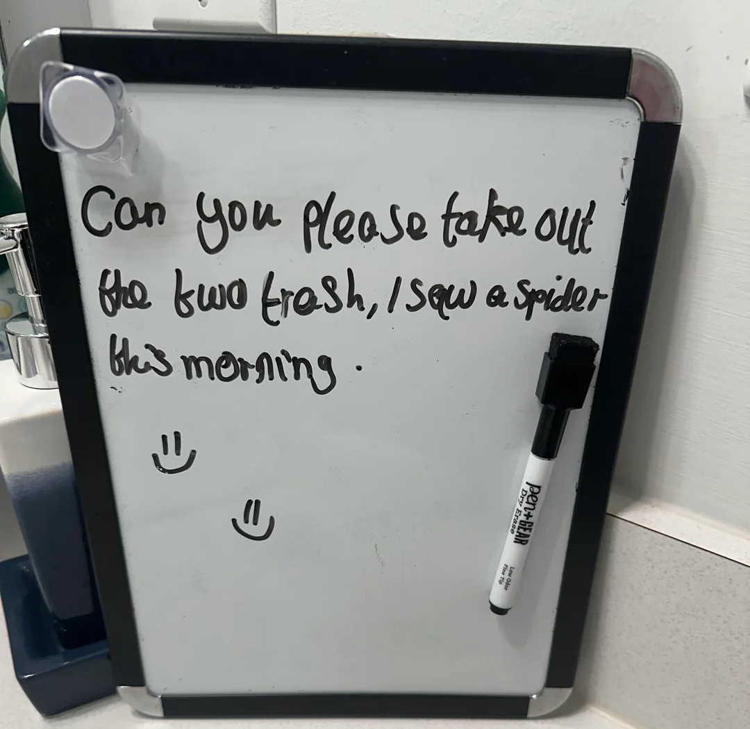 A small whiteboard with a handwritten message: "Can you please take out the bin/trash, I saw a spider this morning." There are also two smiley faces drawn on the board, and a marker is attached to the side.