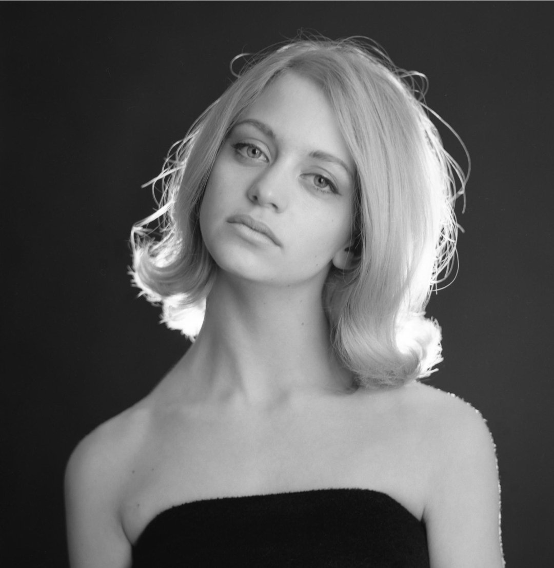 A young woman with light hair styled in soft waves looks at the camera with a neutral expression. She is wearing a strapless black top and is posed against a dark background with backlighting creating a halo effect.