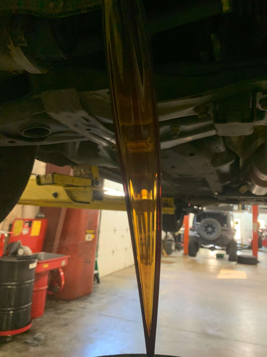 Golden-brown motor oil flows in a steady stream from the underside of a raised vehicle during an oil change in an automotive repair shop.
