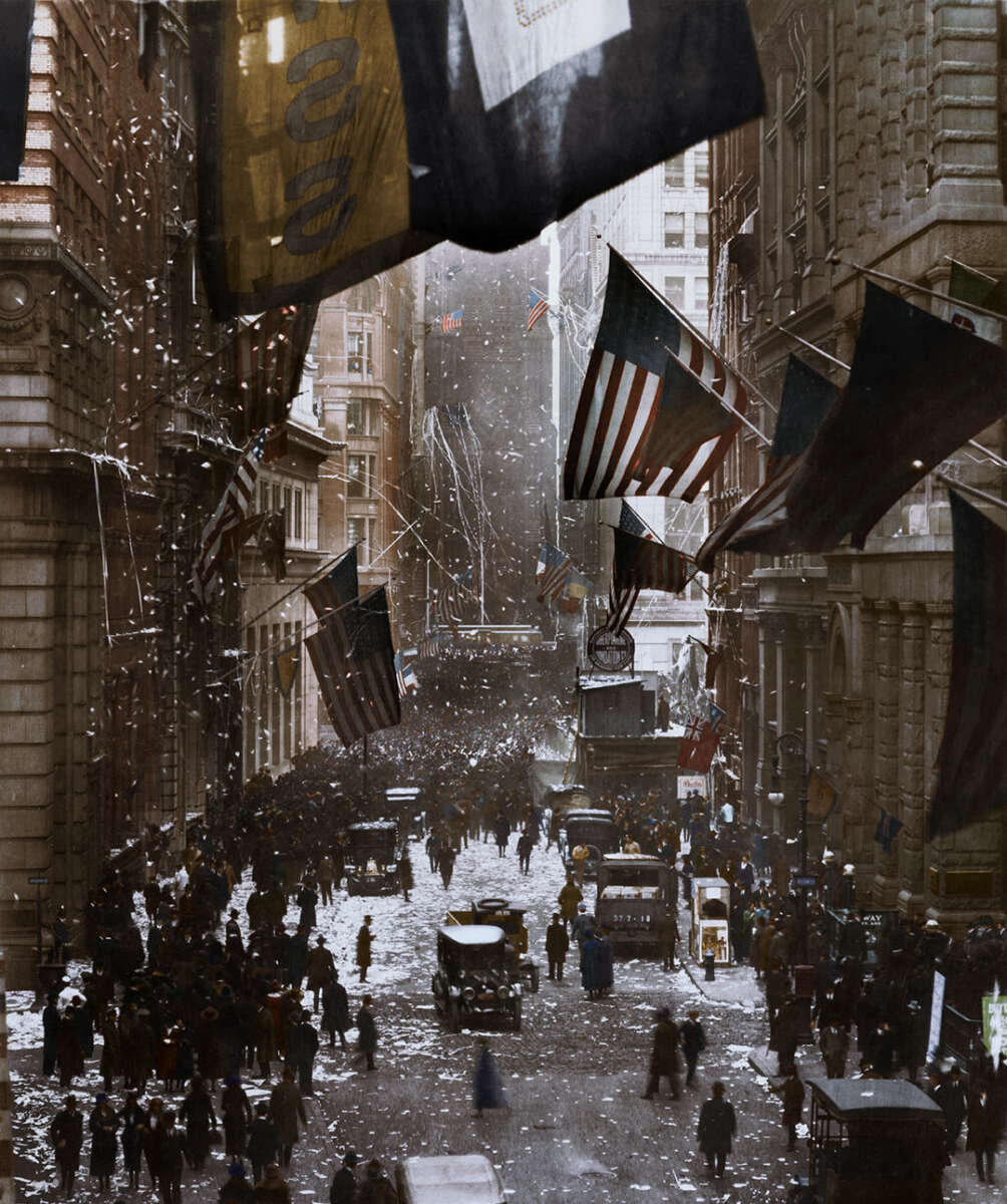 A historic, crowded city street scene with people, old cars, and streetcars. Large American flags hang from buildings, confetti falls, and it appears to be a celebration or parade in early 20th-century New York.