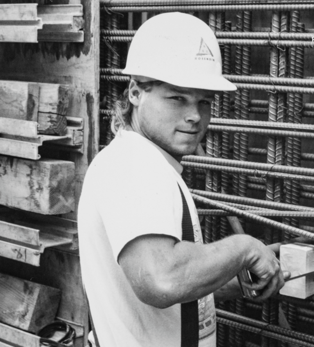 A construction worker in a hard hat and white shirt smiles while working with metal rebar and wooden planks at a building site. The image is black and white.