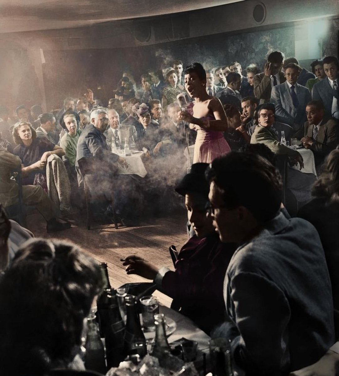 A jazz singer performs on stage in front of a crowded, smoky nightclub filled with people sitting at tables, watching her. The room is dimly lit, with bottles and glasses on the tables.