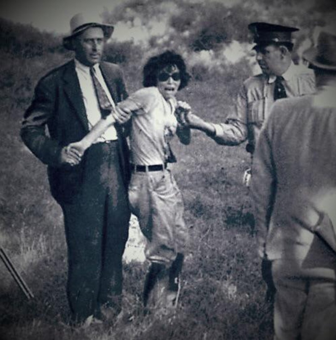 A distressed person wearing goggles is being held by two men, one in a suit and another in a uniform, while a third person in a hat stands nearby on a grassy field.