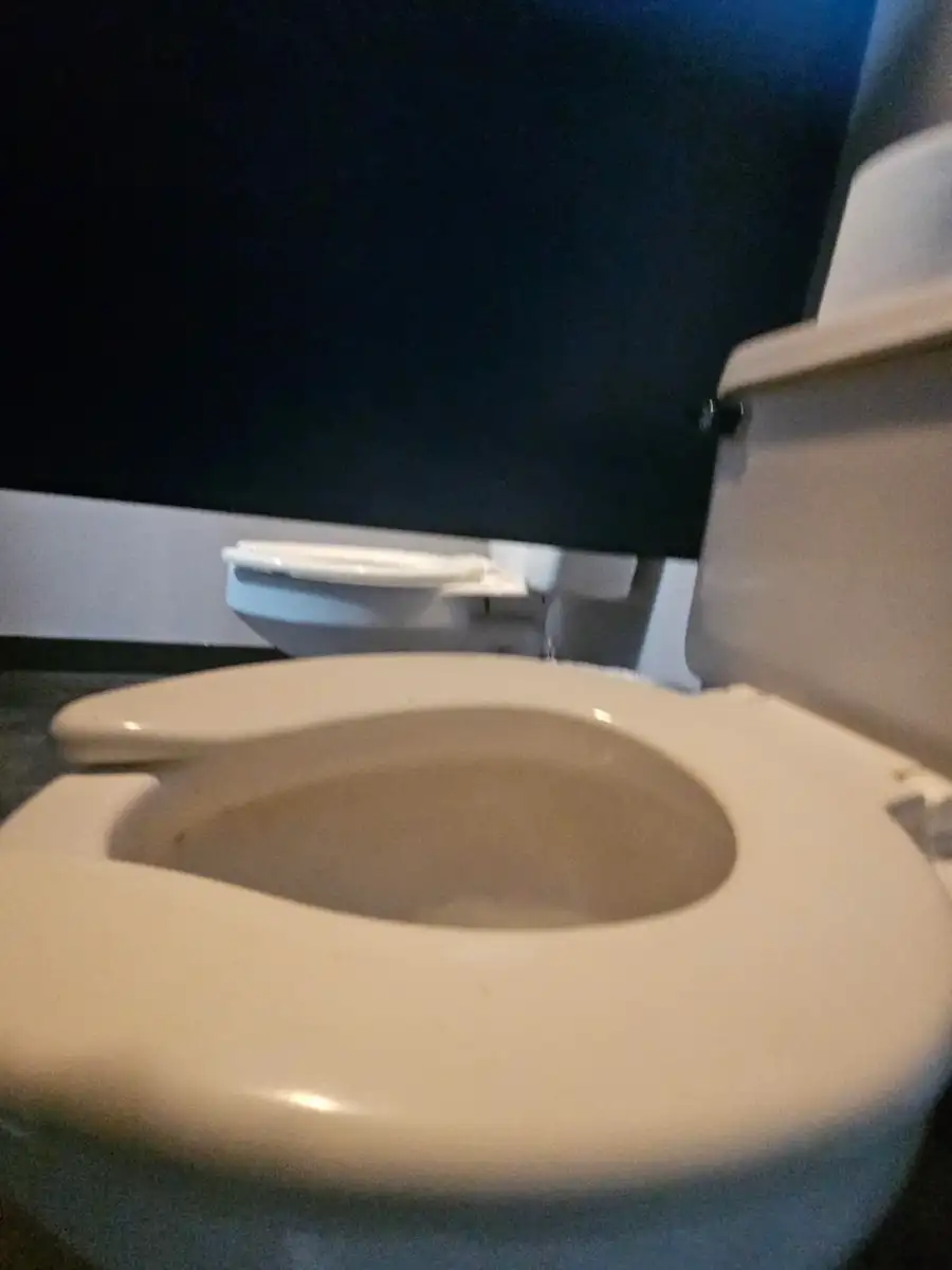 A restroom with two white toilets next to each other, one in the foreground and another in the background, against a dark wall.