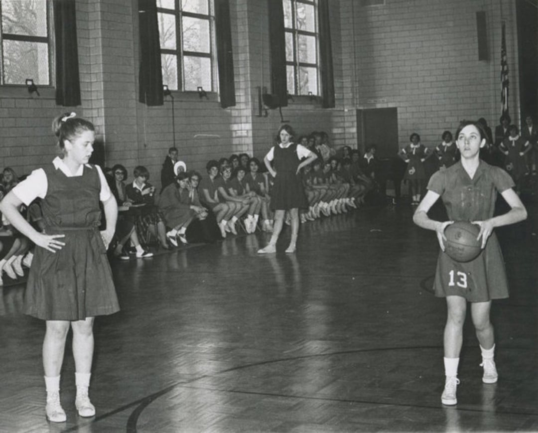 A vintage black-and-white photo of three young women playing basketball in a gym, with one holding the ball. A group of girls in uniform sit along the wall in the background, watching the game.