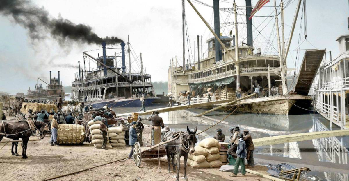 A busy 19th-century river port with steamboats docked, people loading and unloading goods, horses and carts carrying sacks, and smoke rising from a ship, all along a waterfront.