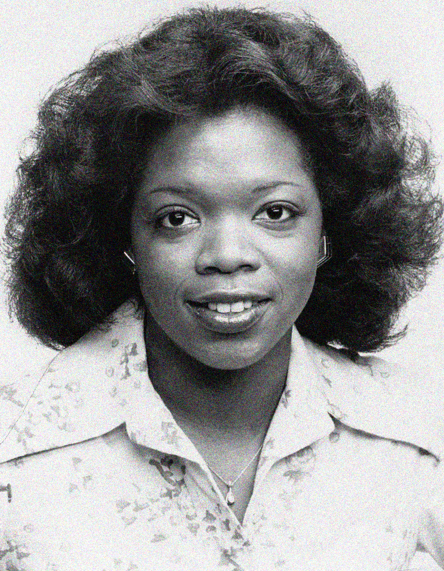 Black and white portrait of a young woman with voluminous, curled hair, wearing a collared floral blouse and a small necklace, looking directly at the camera and smiling softly.