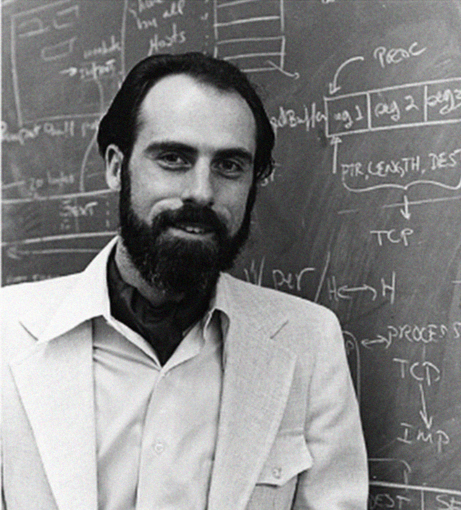 A man with dark hair and a beard, wearing a light-colored suit and scarf, stands in front of a chalkboard filled with technical diagrams and handwritten notes.