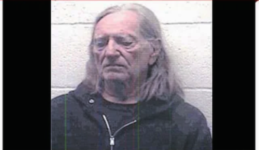 An older man with long gray hair wearing a dark hooded sweatshirt stands against a light-colored cinder block wall, looking downward with a neutral expression.