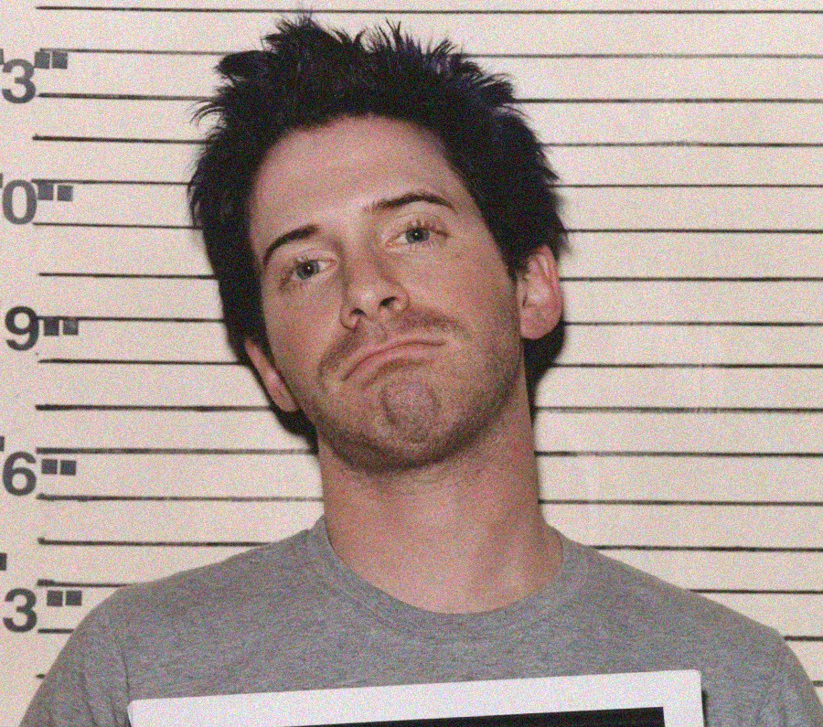 A man with short, dark, messy hair stands in front of a height chart, wearing a gray t-shirt and holding a sign, appearing to have a mugshot taken. He has a neutral expression and is slightly tilted to one side.