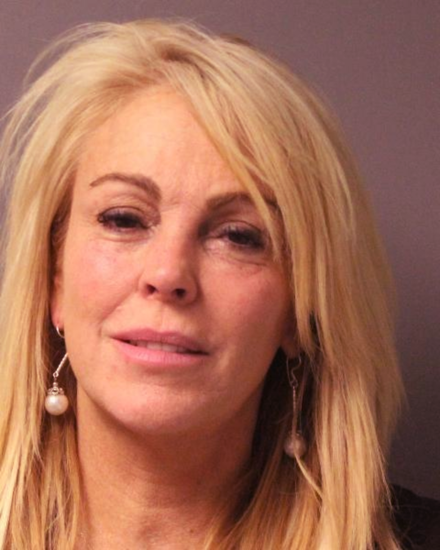 A woman with long blonde hair and light makeup is wearing pearl drop earrings and a black top, standing against a plain background, looking slightly off camera with a neutral expression.