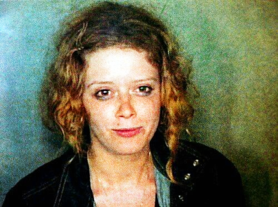 A woman with curly, reddish-blonde hair and pale skin looks at the camera with a neutral expression. She is wearing a dark jacket over a light-colored top, and the background is a muted greenish tone.