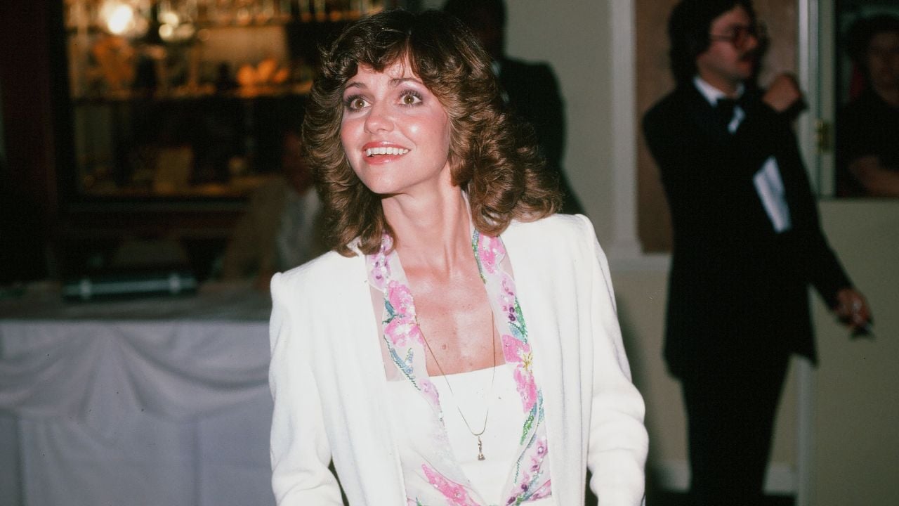 A woman with curly brown hair in a white suit and floral blouse smiles at an event. A man in a tuxedo stands blurred in the background.