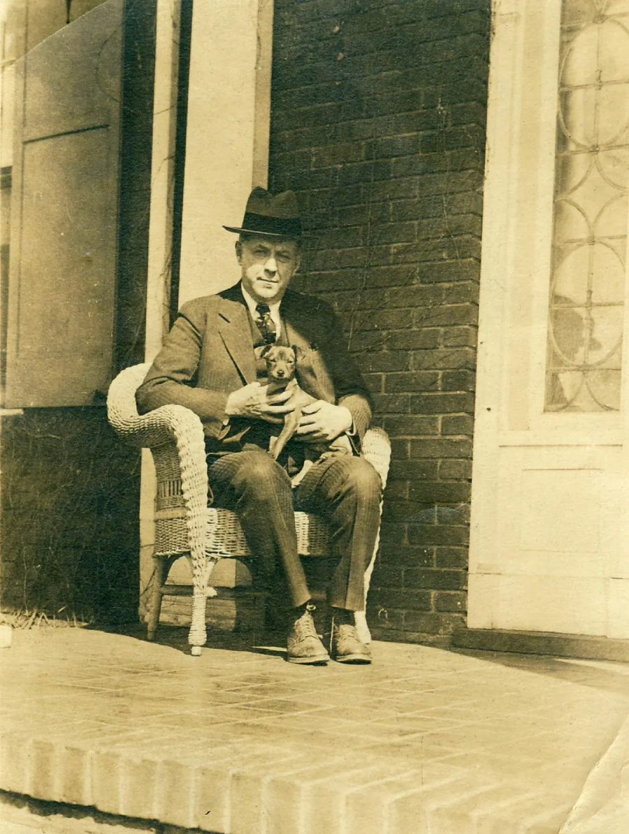 A man in a suit and hat sits on a wicker chair on a porch, holding a small dog in his lap. The setting appears to be outside a brick building with a door and window nearby. The photo is sepia-toned.