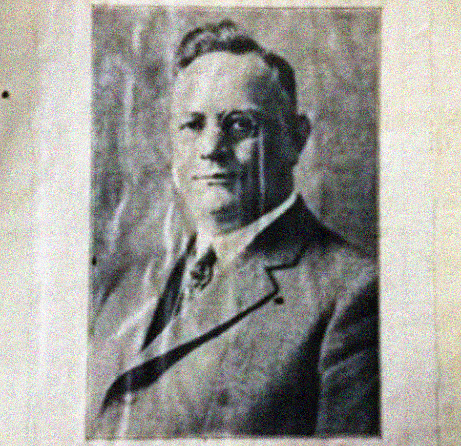 A black and white, vintage portrait photo of a man in a suit and tie, posing formally and facing the camera. The image appears old and slightly worn or faded.