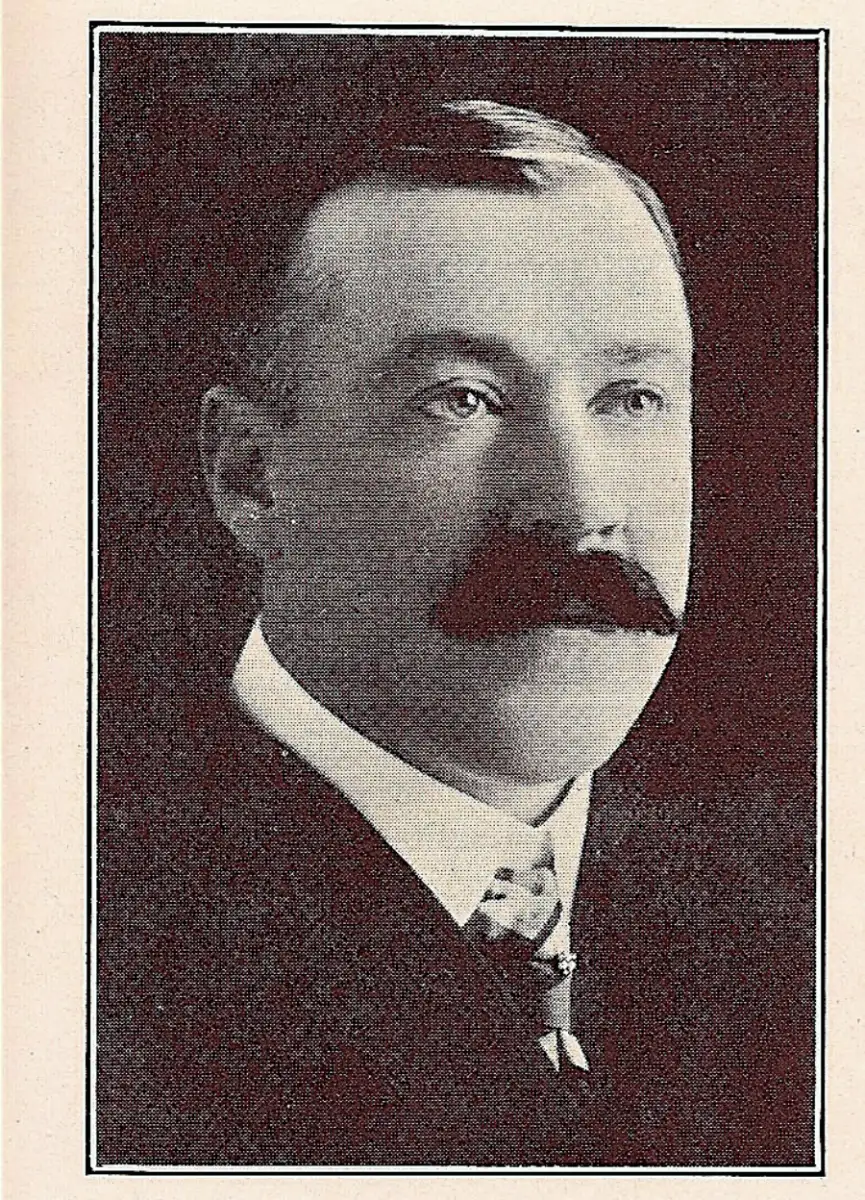 A sepia-toned portrait of a man with a large, dark mustache, wearing a suit and tie, looking slightly to his right. The image has a vintage, early 20th-century style.