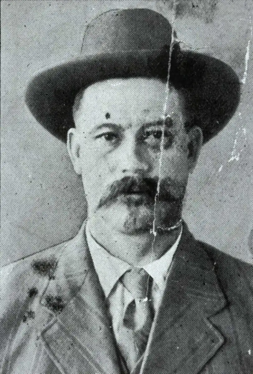 Black and white portrait of a man with a mustache wearing a wide-brimmed hat, suit, and tie, looking directly at the camera. The photo shows visible scratches and signs of age.