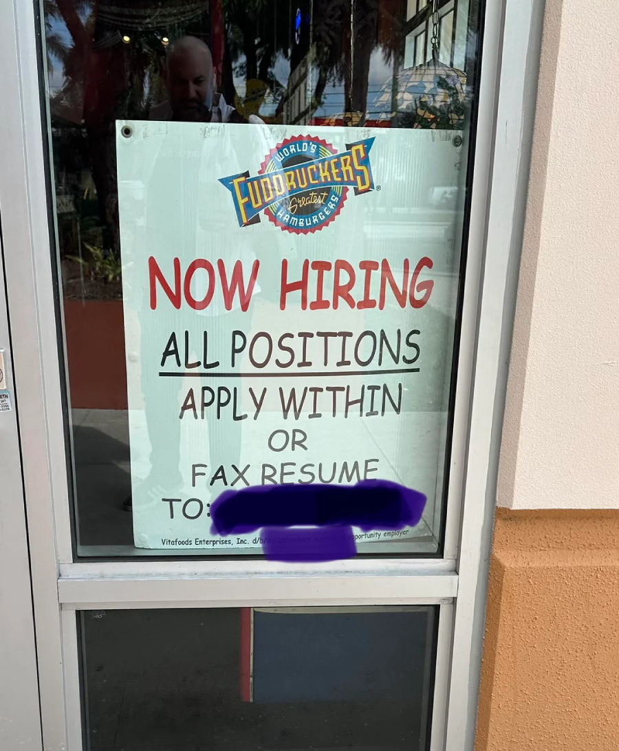 A sign on a glass door advertises that Fuddruckers is now hiring for all positions, asking applicants to apply within or fax a resume to a number that is partially obscured.