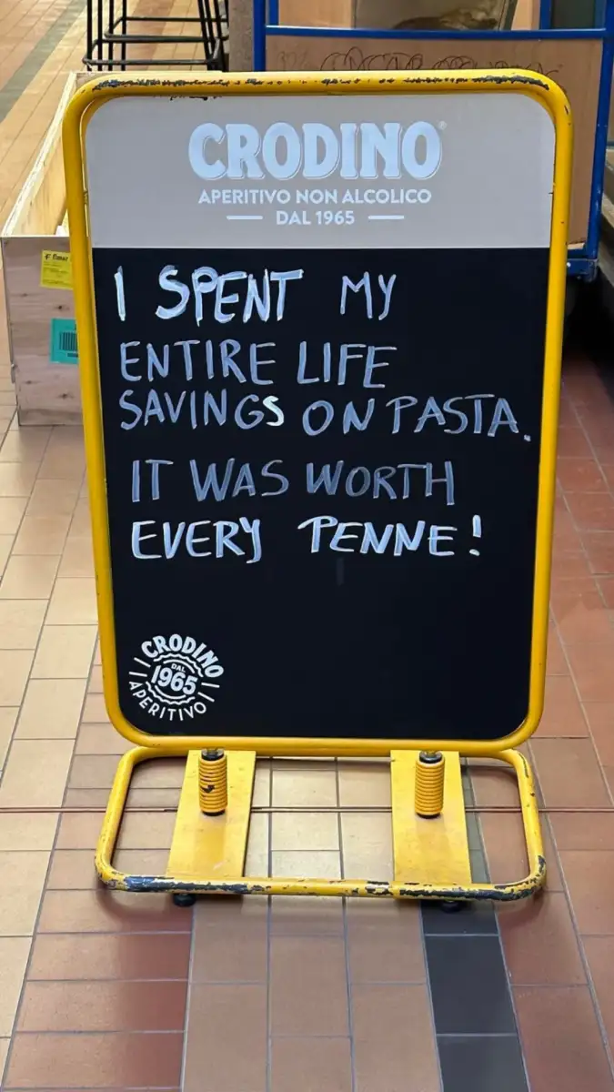 A blackboard sign with the text: "I spent my entire life savings on pasta. It was worth every penne!" written in white chalk. The sign is framed in yellow and placed on a tiled floor.