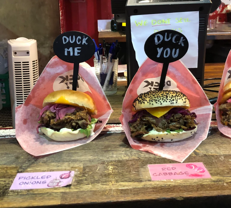Two burgers labeled “Duck Me” and “Duck You,” each with toppings like pickled onions and red cabbage, are displayed on a counter, with handwritten signs and a menu in the background.