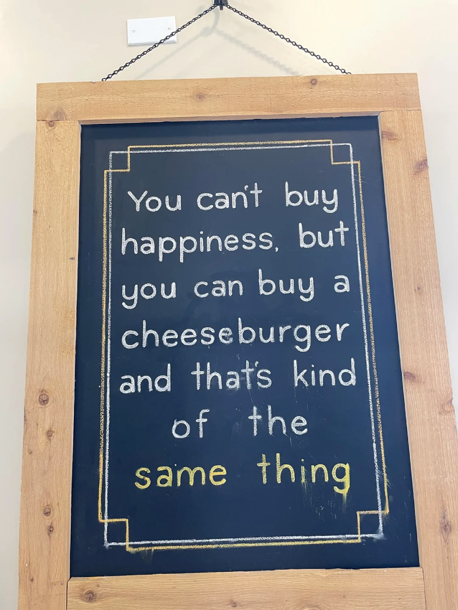 A chalkboard sign in a wooden frame reads: "You can't buy happiness, but you can buy a cheeseburger and that's kind of the same thing," with "same thing" highlighted in yellow.